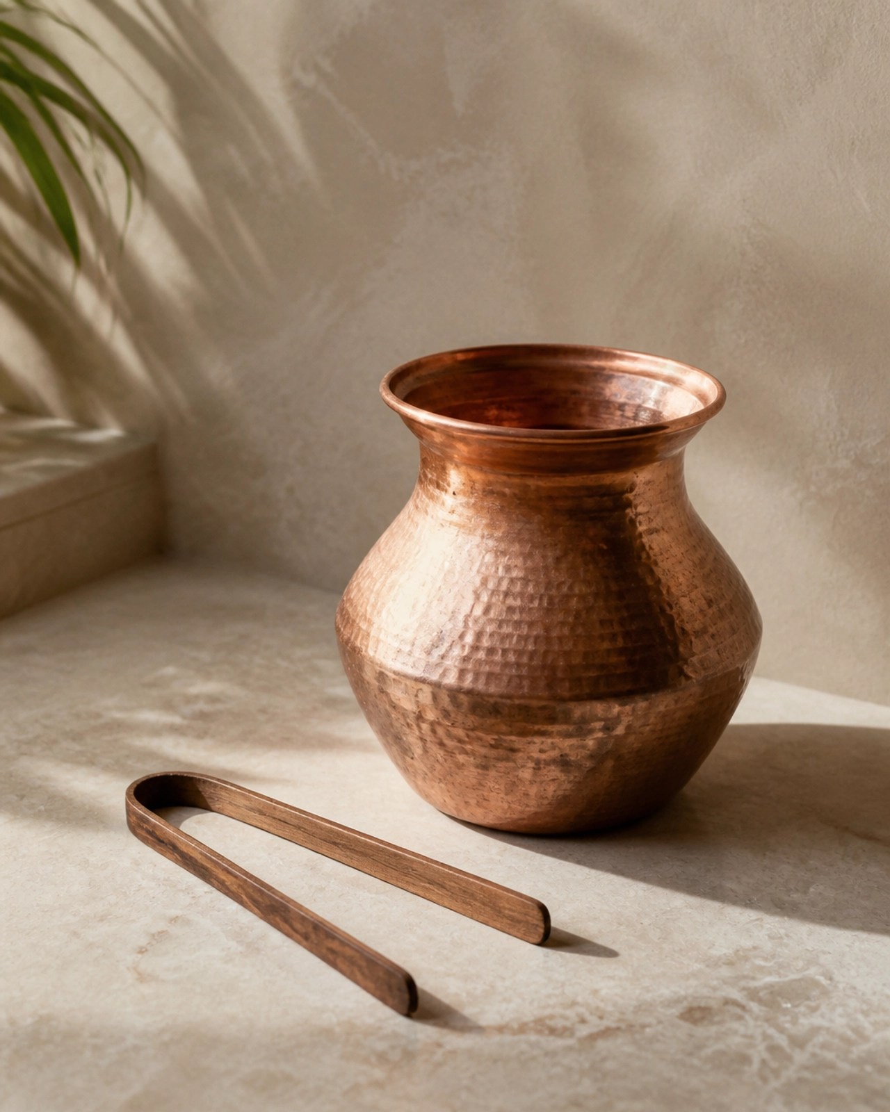 A copper water vessel and a wooden tongue scraper resting on a light beige stone surface in soft lighting