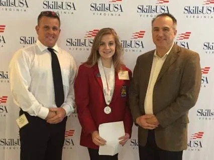 A student in a red SkillsUSA blazer stands proudly between two industry professionals, highlighting the connection between education and industry.