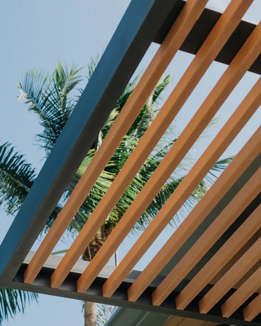 A close-up of a custom louvered roof system featuring premium aluminum frames and warm wood-toned slats, demonstrating the blend of natural aesthetics with modern durability against a tropical sky.