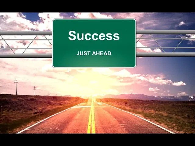 A highway sign displaying Success JUST AHEAD over a sunlit road