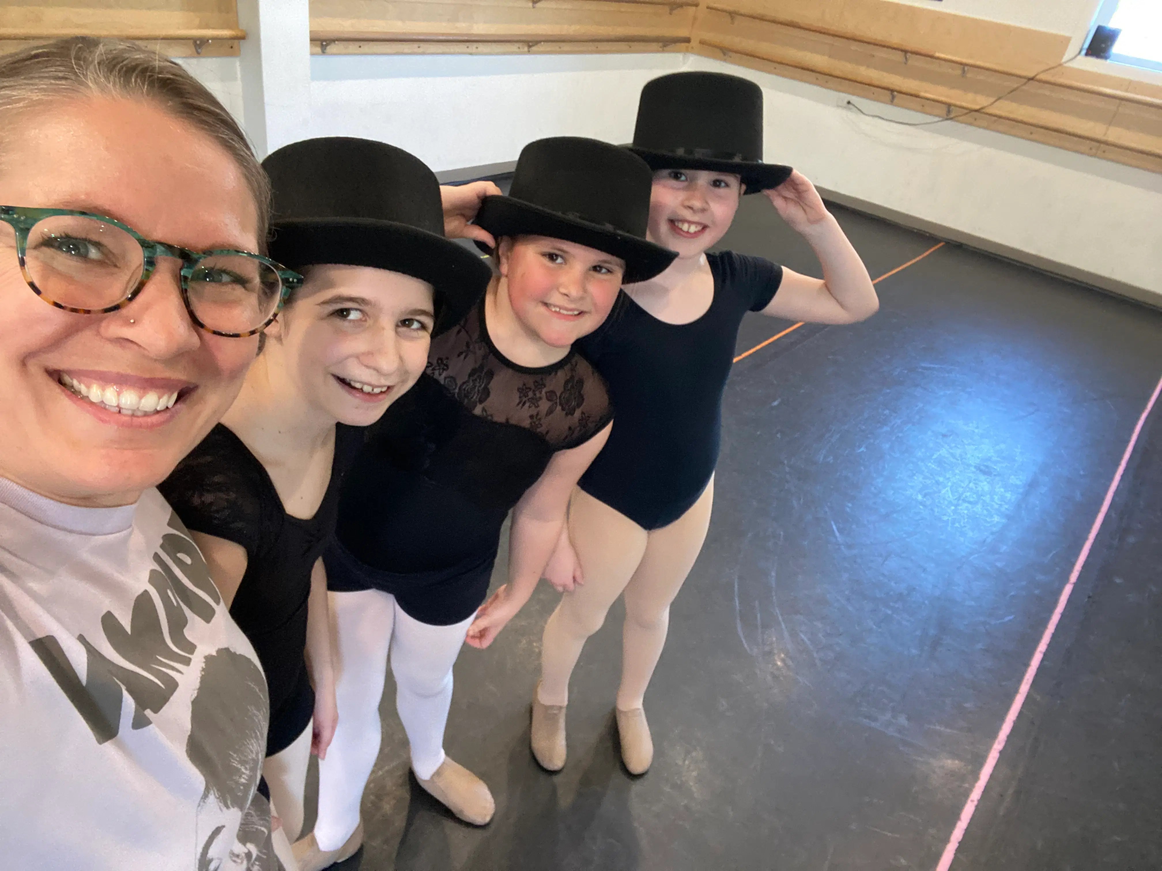 Three young dancers in black leotards, tan tights, and jazz shoes pose with top hats alongside their instructor in a warm, supportive studio setting.