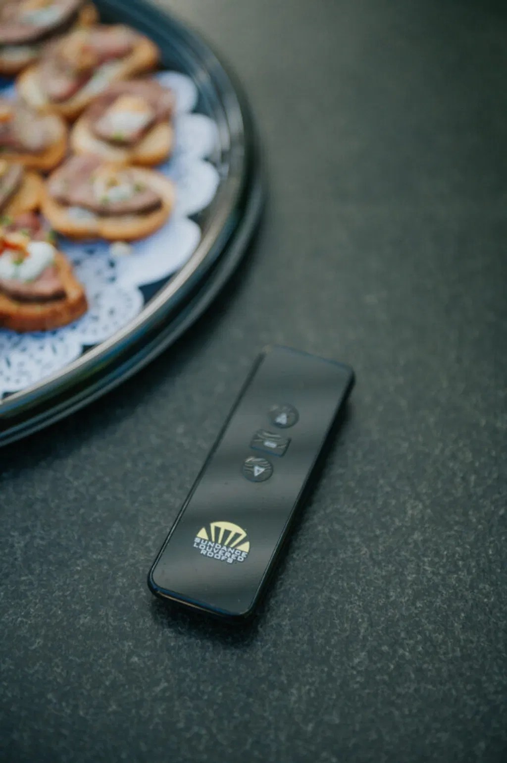 A motorized pergola remote beside gourmet appetizers, representing simple control during an outdoor gathering