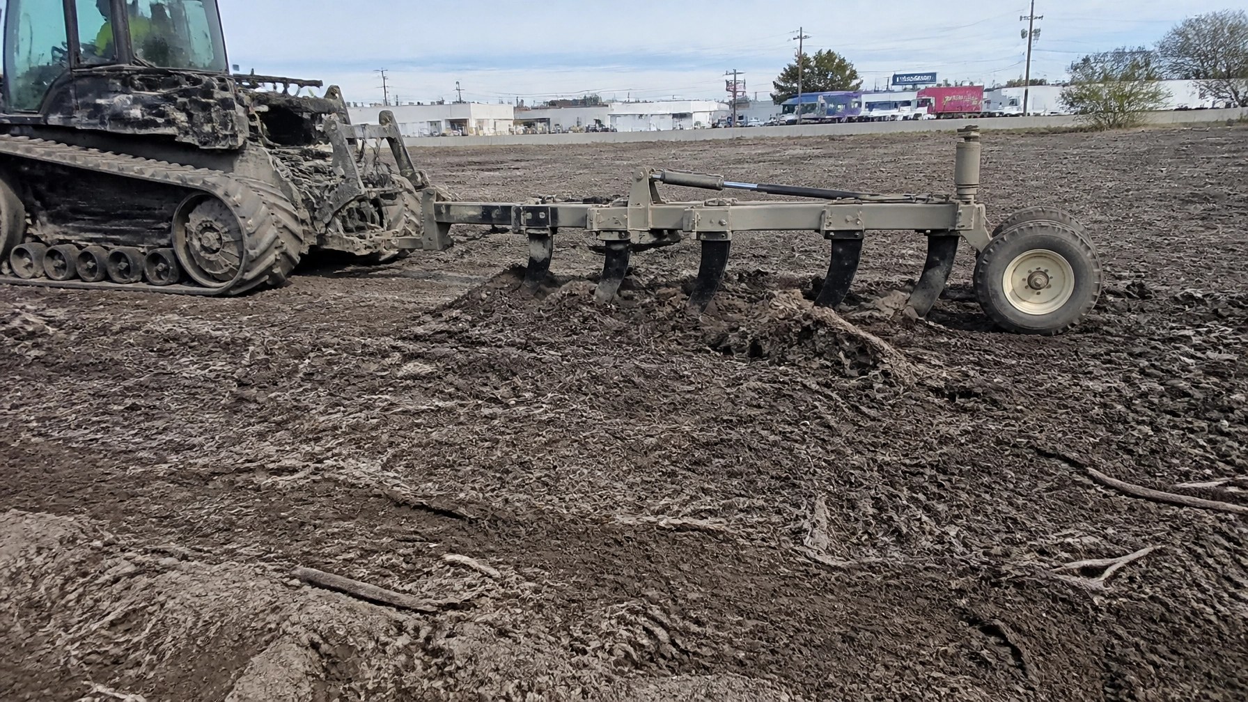 Authentic daylight field photo of brand-neutral equipment performing subsoil restoration on a commercial site. Real dirt, mulch, clay, and track marks are visible. The framing is natural and slightly imperfect, like a crew member captured it on-site with a phone or DSLR. No logos. No CGI look.