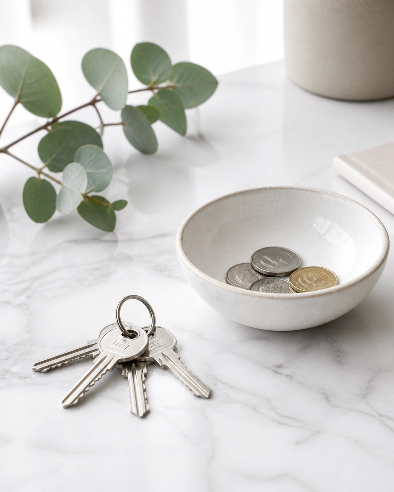 A minimalist close-up of silver house keys next to a ceramic bowl with coins on a white marble surface.