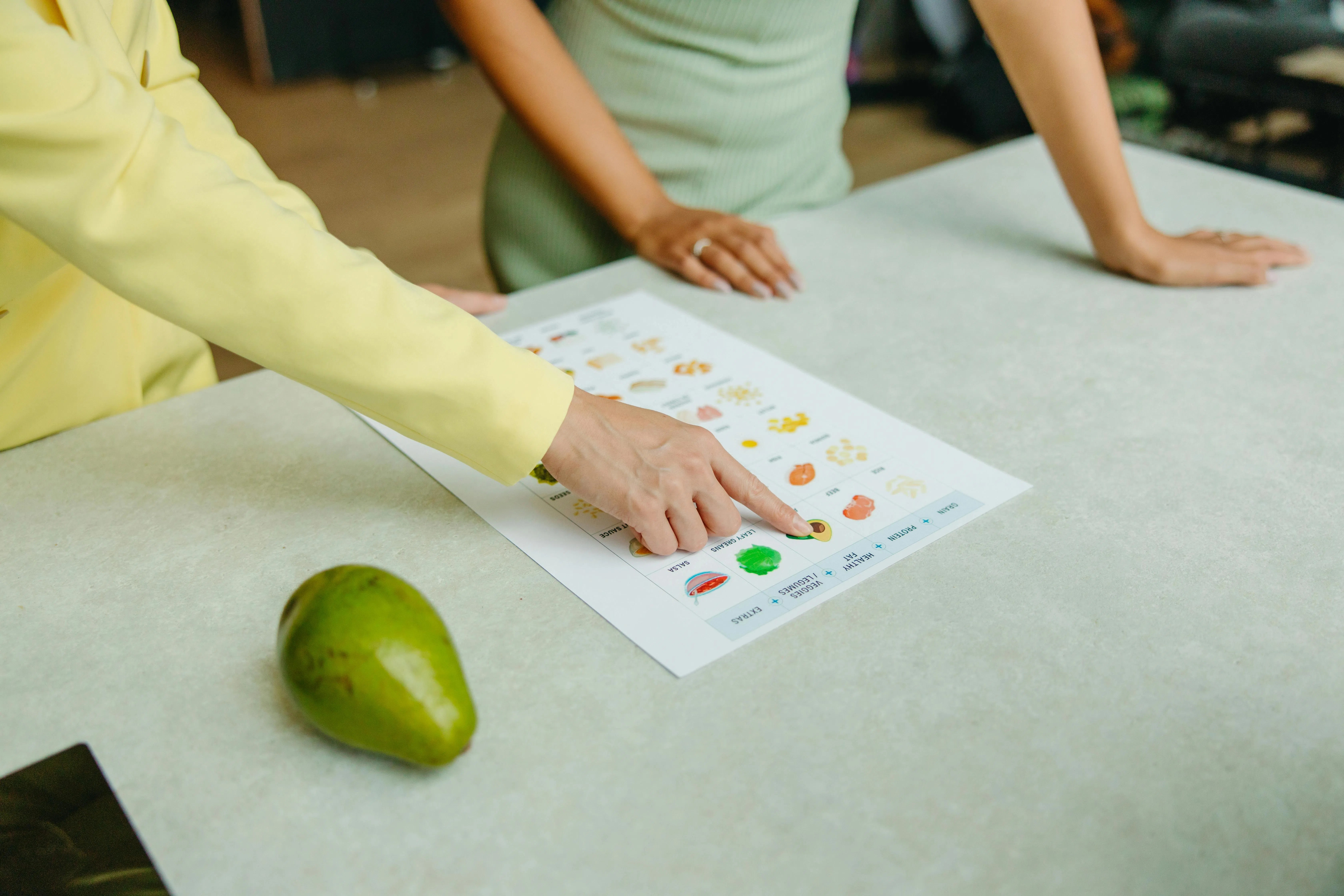 A registered dietitian points to a nutrition chart on a table, providing personalized education to a client. An avocado sits nearby, highlighting practical meal planning.
