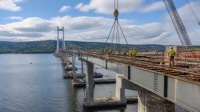 A high-resolution editorial photograph of a major highway bridge construction project over the Hudson River in Upstate New York, showing steel girders, cranes, and workers in safety gear under dramatic natural light.