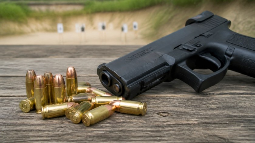 A close-up realistic photo of 9mm Luger full metal jacket cartridges next to a modern handgun on a wooden shooting bench at an outdoor range, natural lighting