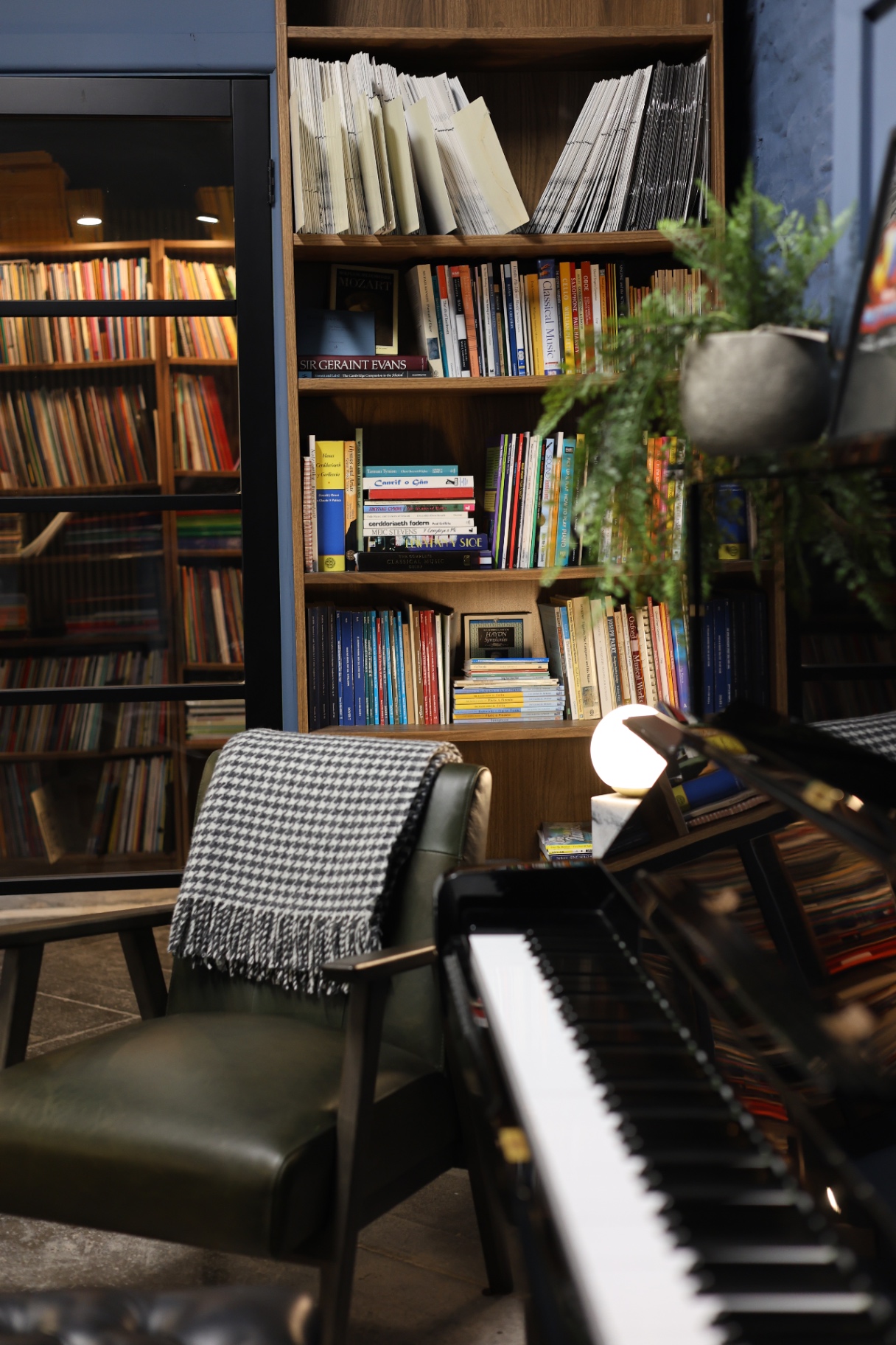 AberMusic Studio Corner A modern piano, comfortable armchair with a houndstooth throw, and well-stocked bookshelves filled with sheet music, music theory books, and learning resources. Designed to inspire lessons with conservatoire-trained teachers in a creative environment.