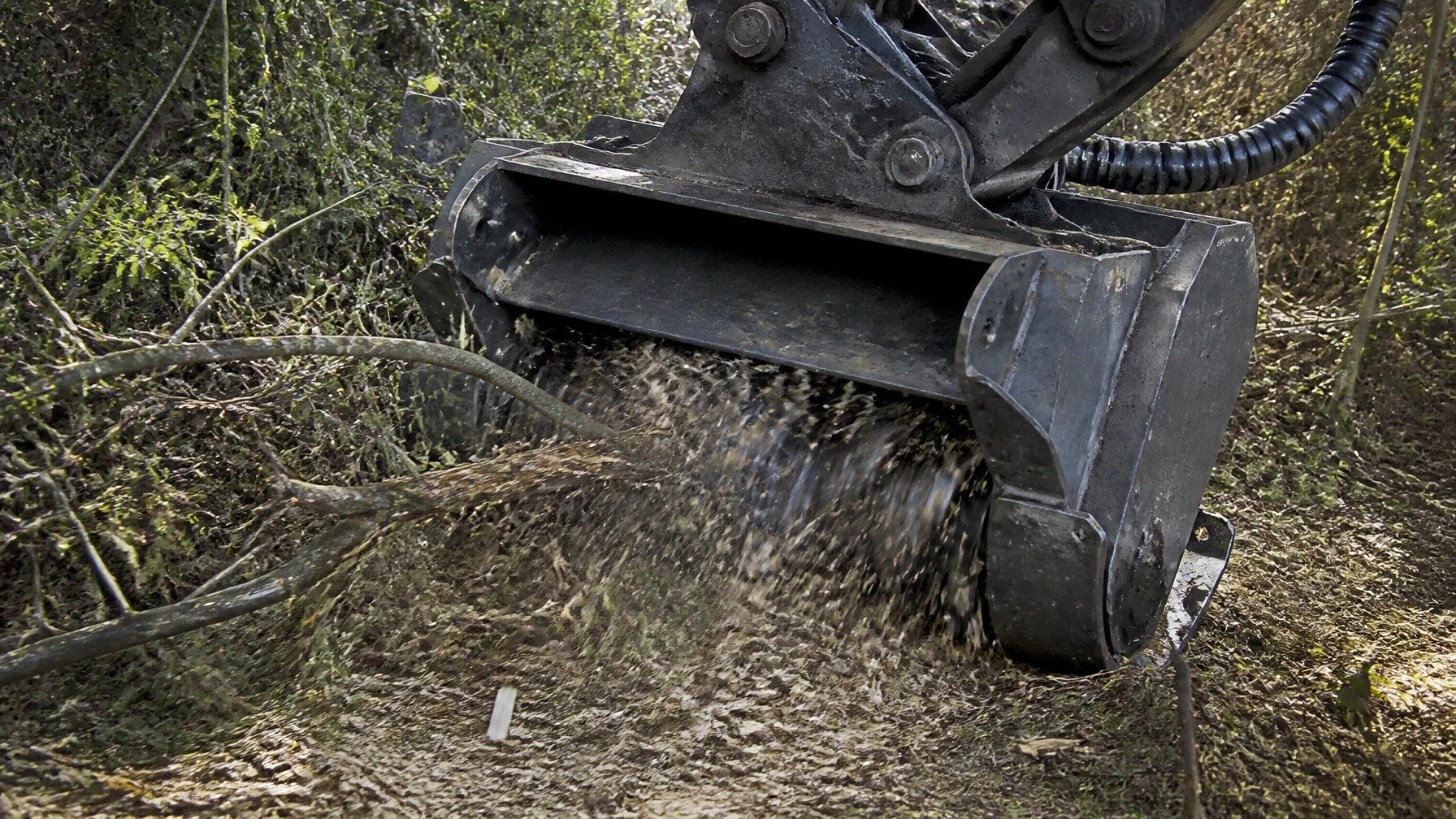 Authentic high-action field photo of a brand-neutral forestry mulcher engaging dense brush on a commercial jobsite in daylight. Wood chips, debris, dirt, and mulch are visible in a raw, handheld documentary-style capture with natural light and imperfect field composition.