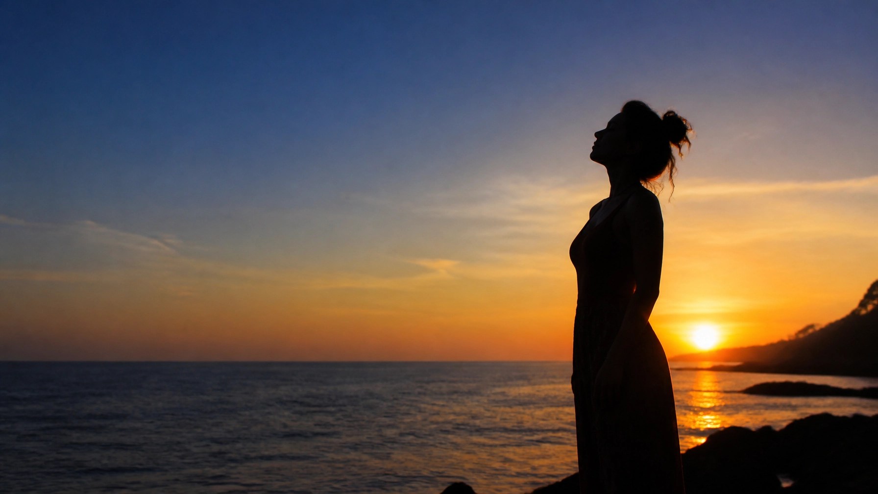 A woman standing peacefully by the ocean at twilight in silhouette