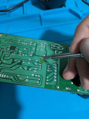 Technician performing a precision soldering repair on a green printed circuit board.