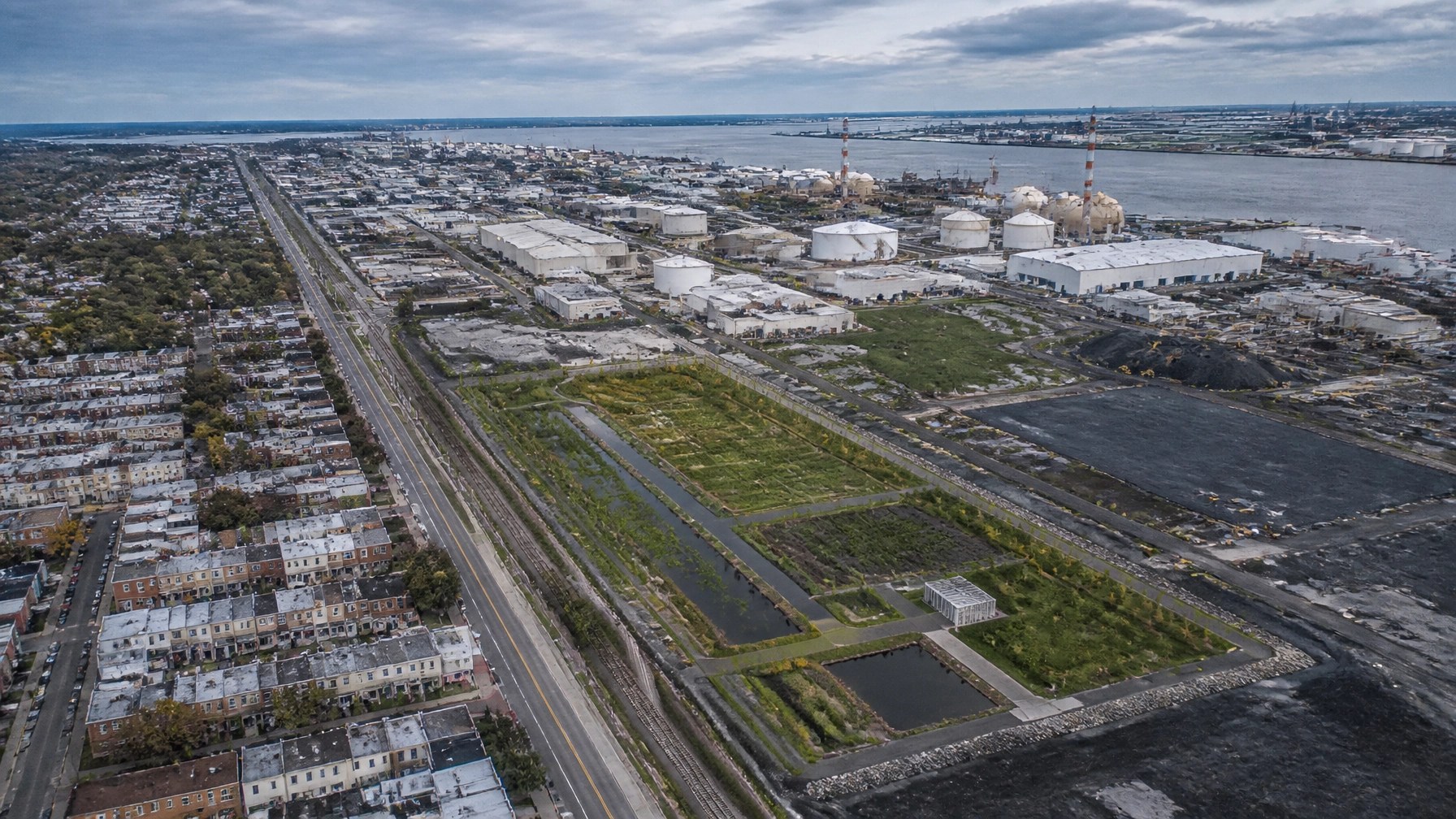 Aerial view of an urban industrial corridor beside a dense residential neighborhood, with a redevelopment parcel and stormwater buffer