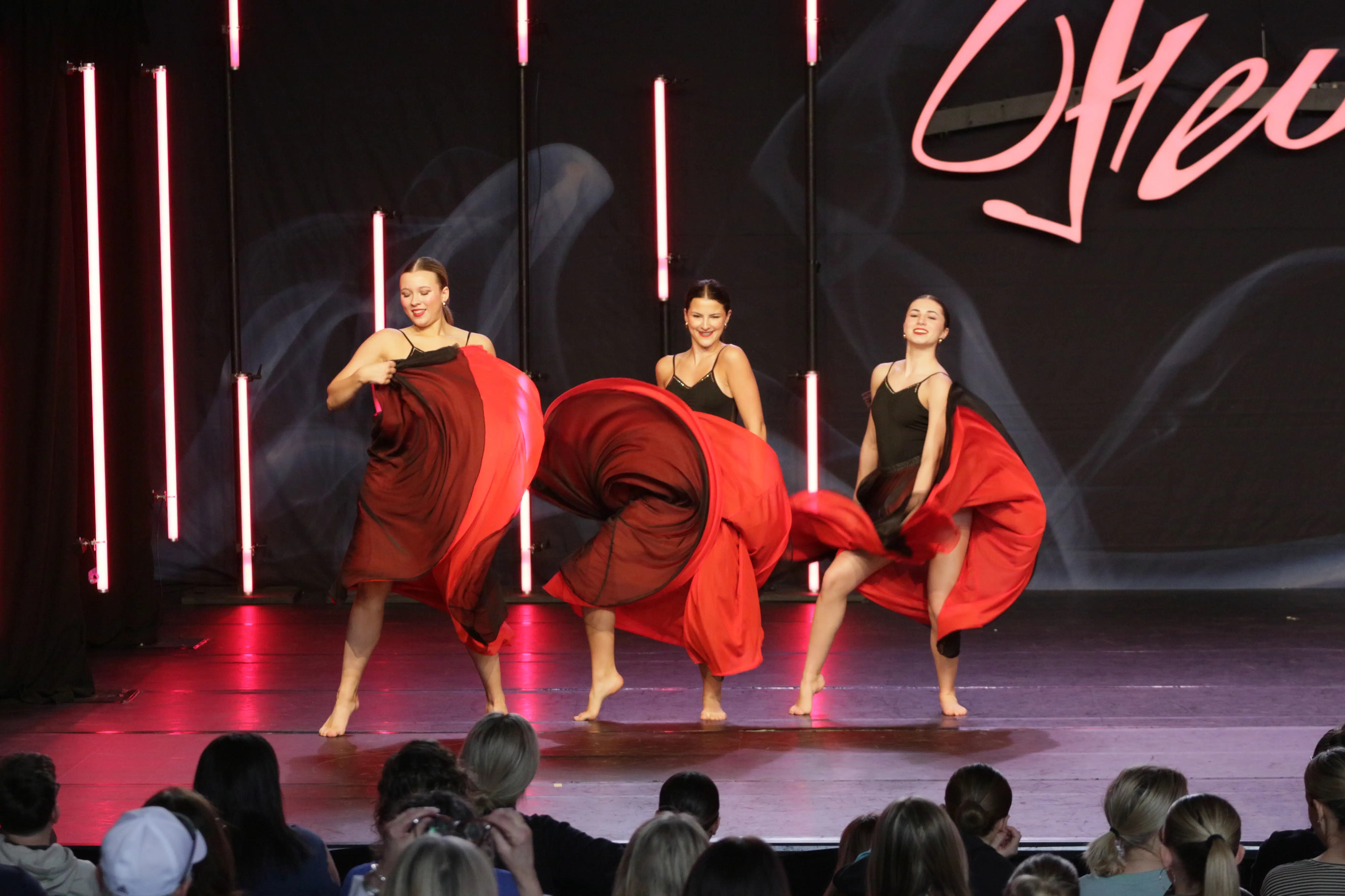 Three dancers perform together on stage in black and red costumes with flowing skirts, showing teamwork and performance power.