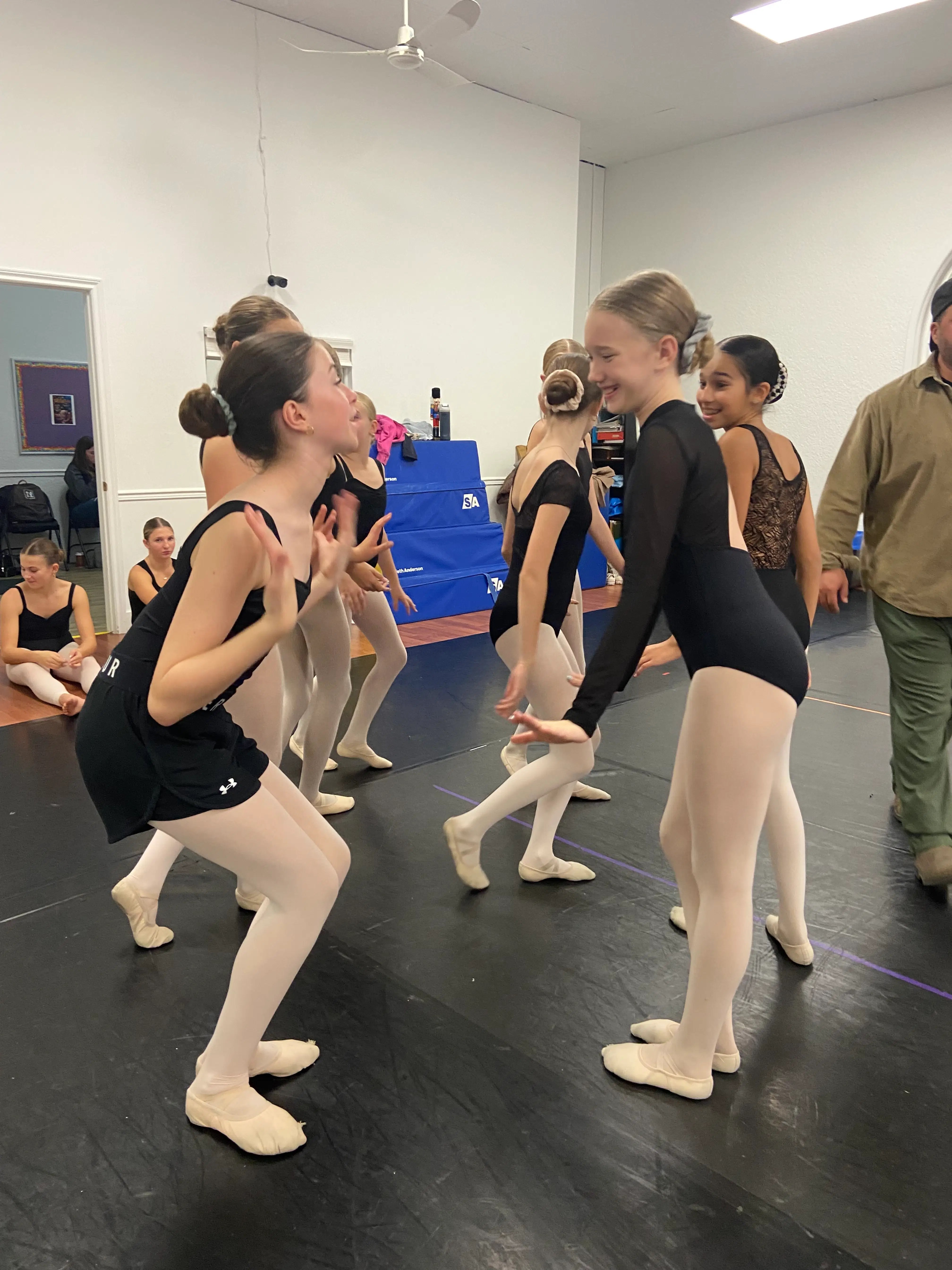 A group of 8-10 year old dancers in black leotards laugh and interact together on the studio floor, showing the connection and focus that grow inside Intensive Study training.