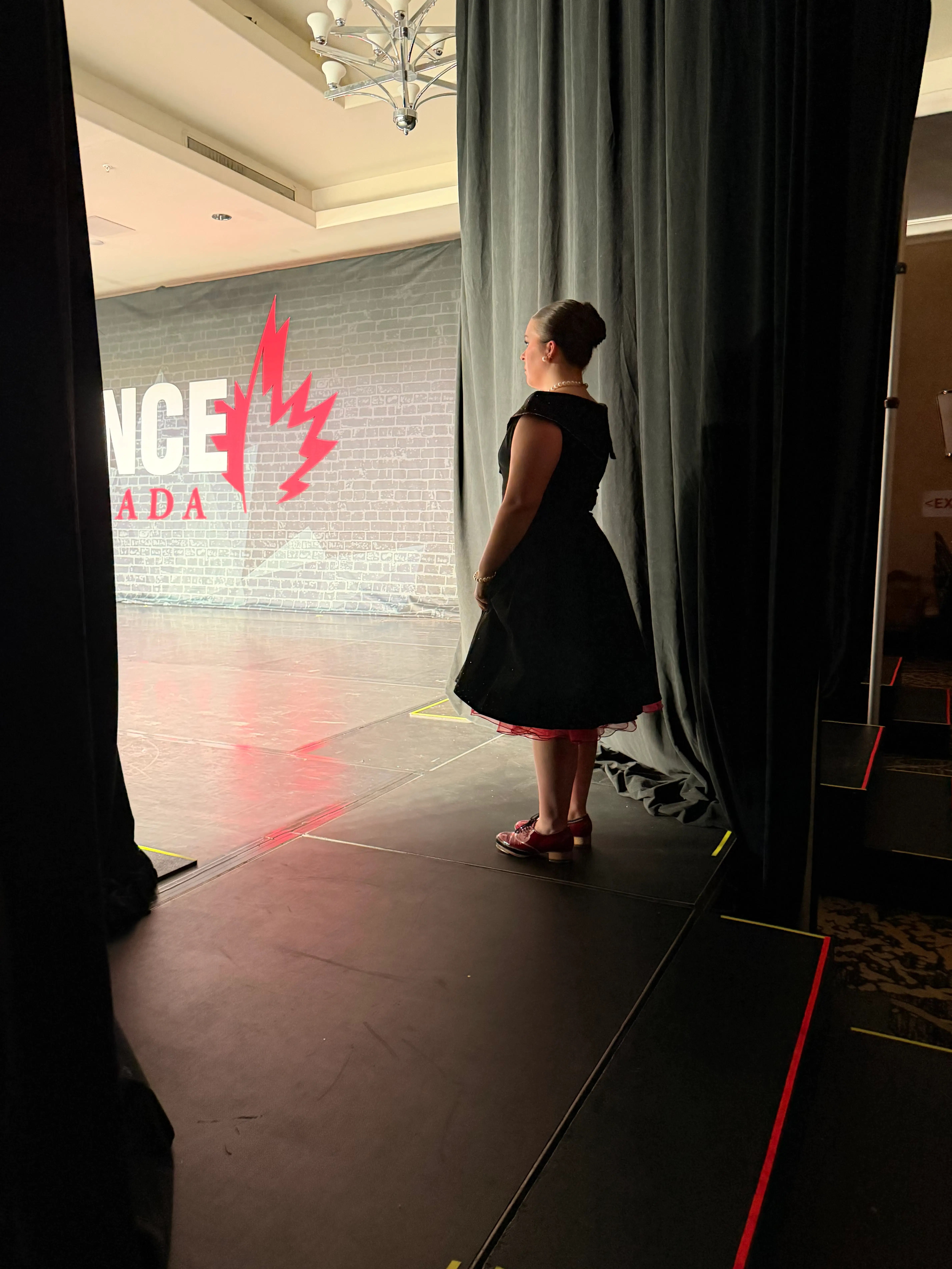 A dancer waits backstage in a black dress, preparing to go on stage under the lights.