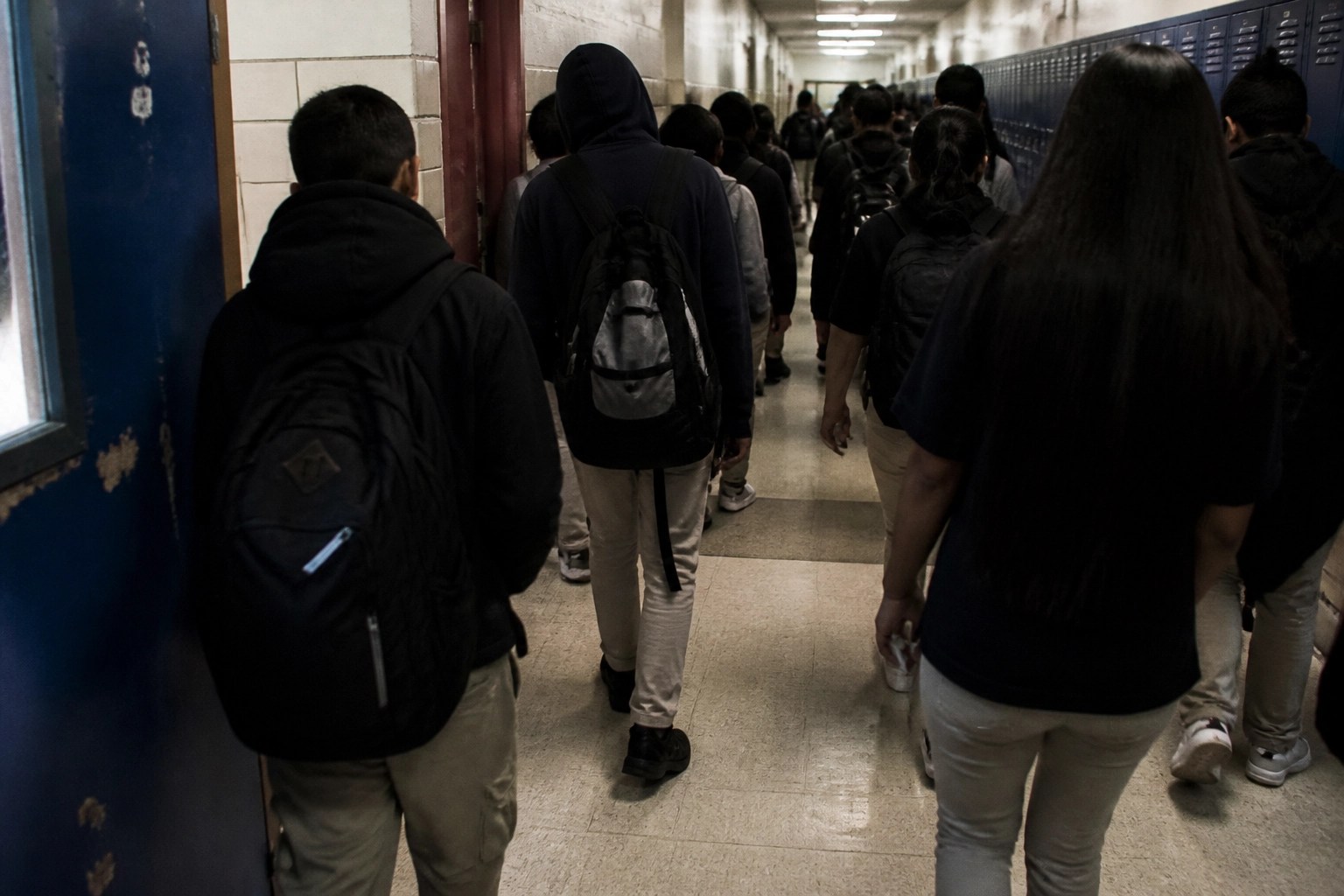Crowded NYC public school hallway with gritty lockers and teenagers moving through the pressure of the day, faces obscured.