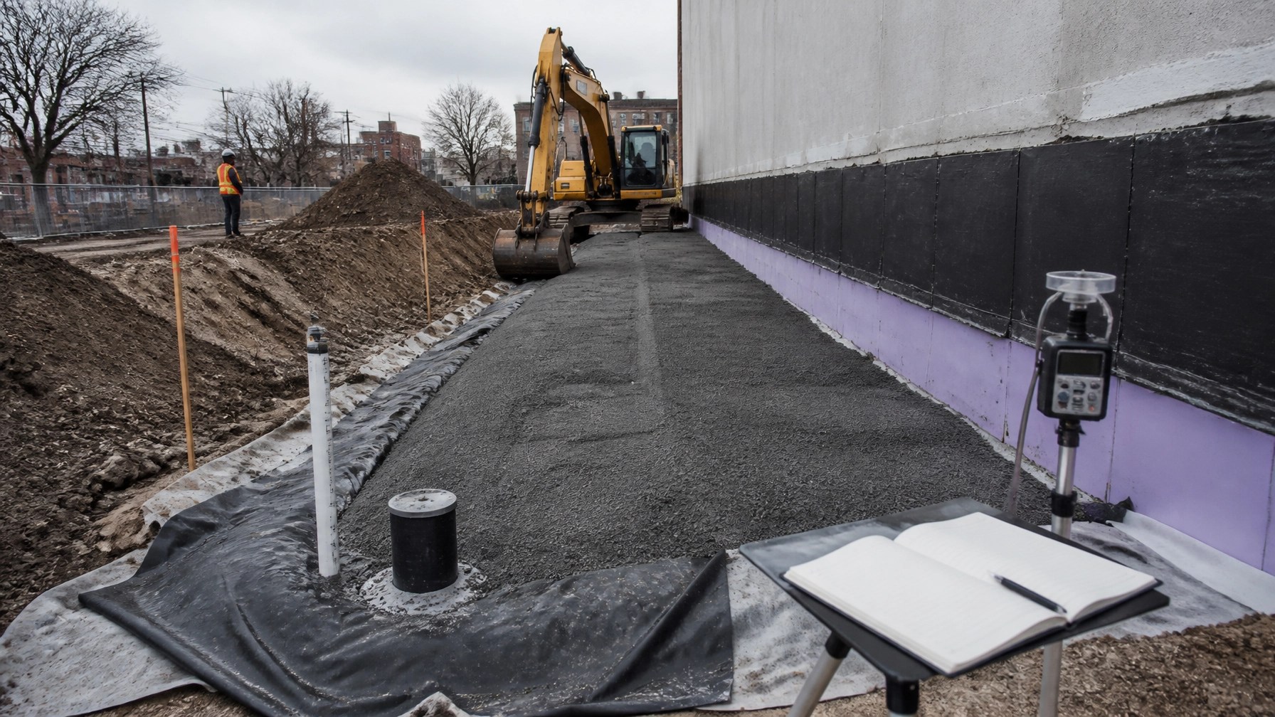NYC brownfield excavation with engineered vapor barrier, monitoring equipment, survey stakes, and field oversight