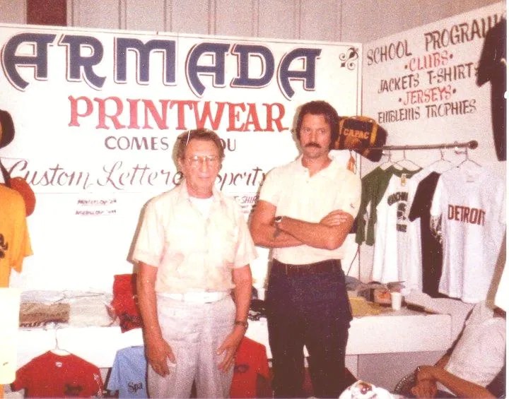 The founders of API Corporate at their original printwear booth, showcasing their deep roots in the screen printing industry.