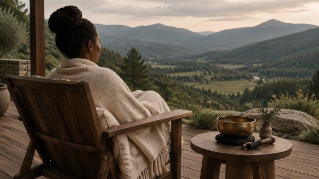 A person of color wrapped in a cream throw sits on a wooden chair overlooking a muted green mountain valley, with a small bowl resting on a nearby table