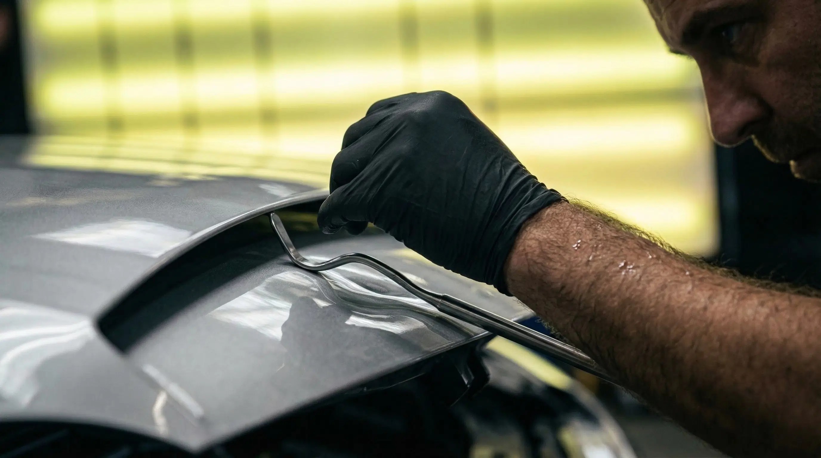 An elite PDR technician wearing black gloves precisely manipulates a specialized tool under a vehicle panel, using high-focus lighting to identify surface imperfections.