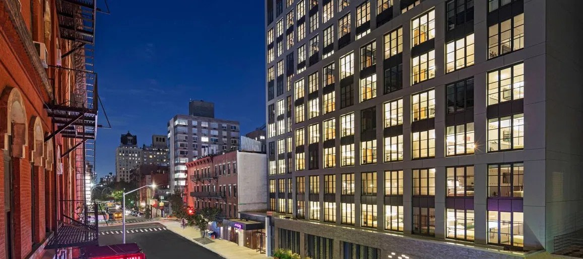 Mixed-use mid-rise building in a densely developed NYC neighborhood at dusk.