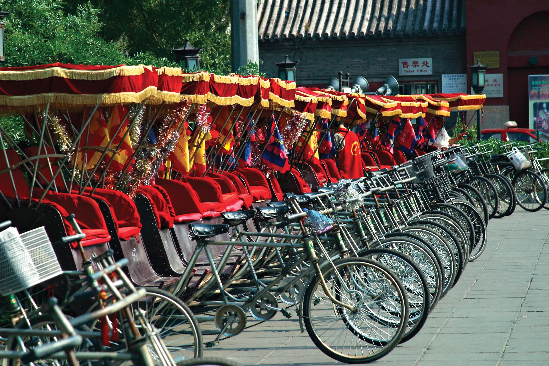 A line of parked bicycle rickshaws with red seats and canopies.