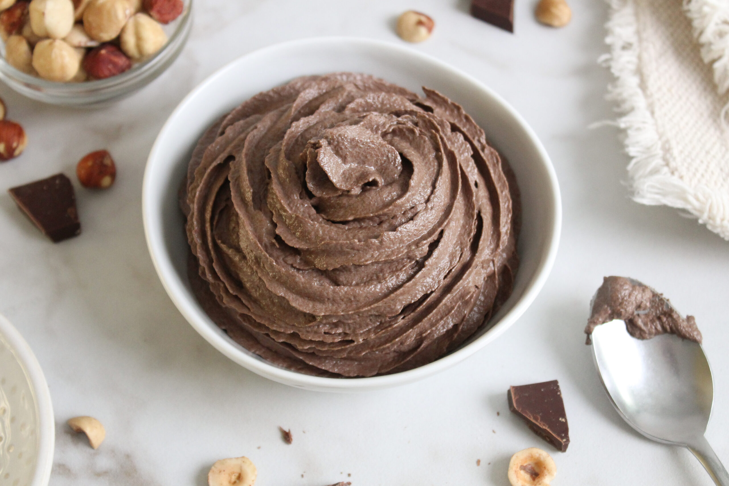 Chocolate Hazelnut Nutella Buttercream Frosting in a bowl with chocolate pieces and hazelnuts around
