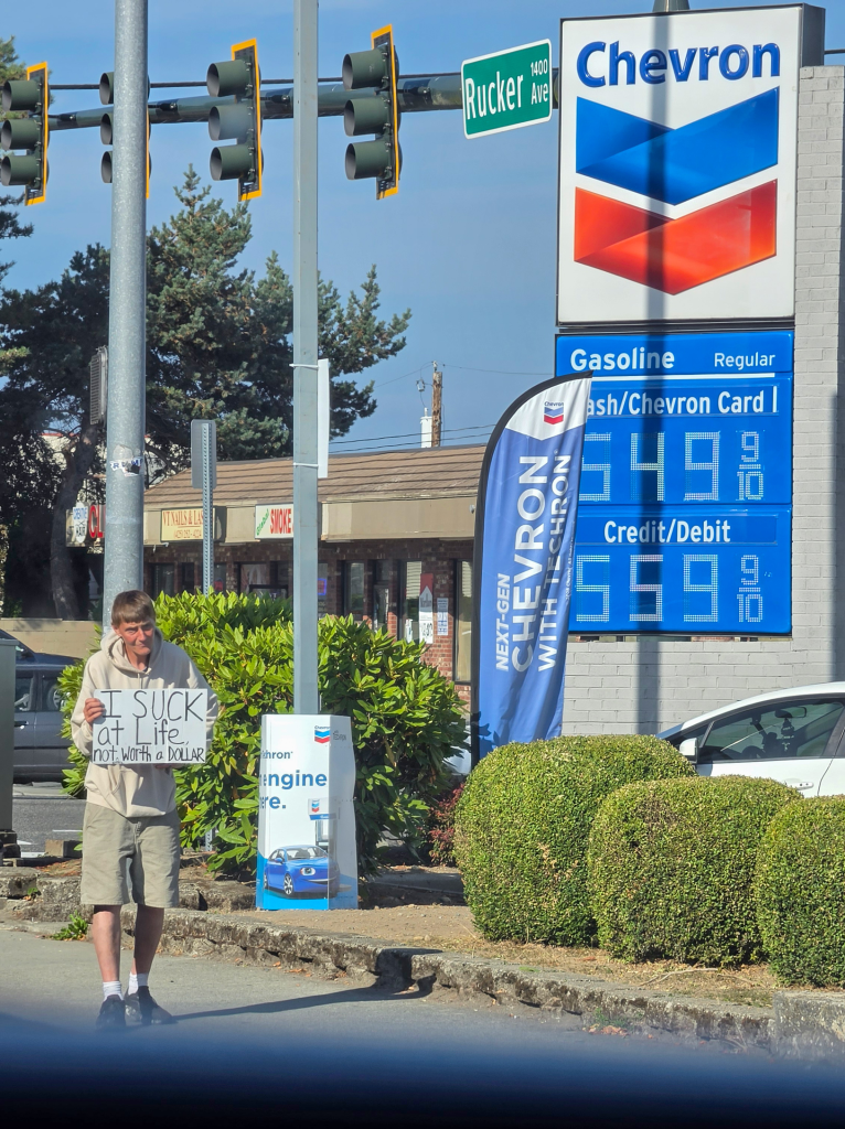 Man in Everett holds a sign reading “I suck at life, not worth a dollar” beside a Chevron sign advertising .49 gas. The 20-minute gallon