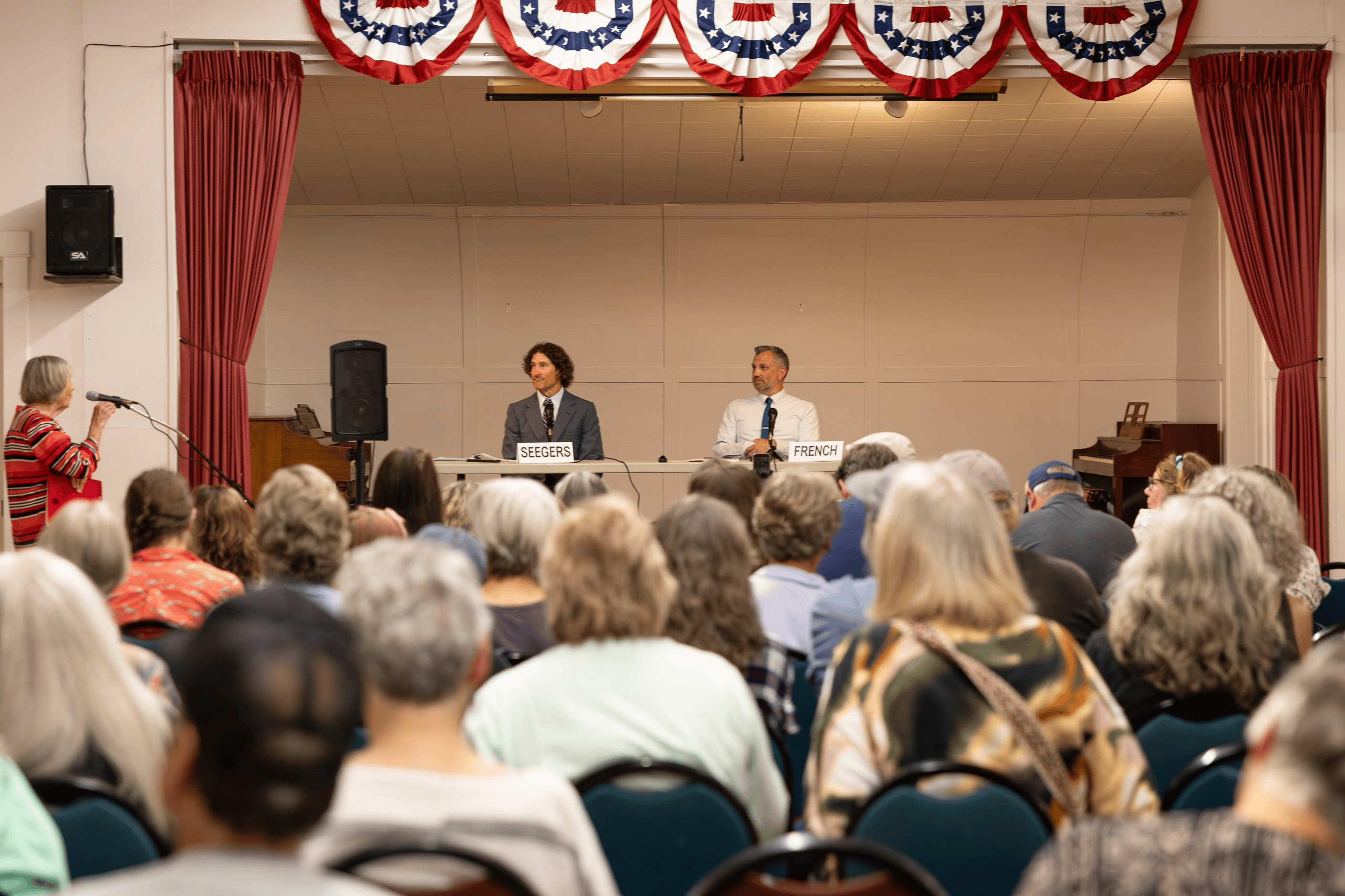 Jake Seegers and Mike French listen from the stage as an attendee asks a question before a crowded audience. Clallam County District 3 commissioner candidates Jake Seegers and Mike French seated at a table facing a full room at the Sequim Prairie Grange.