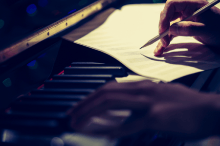 one hand on a piano keyboard, the other holding a pencil over music manuscript paper