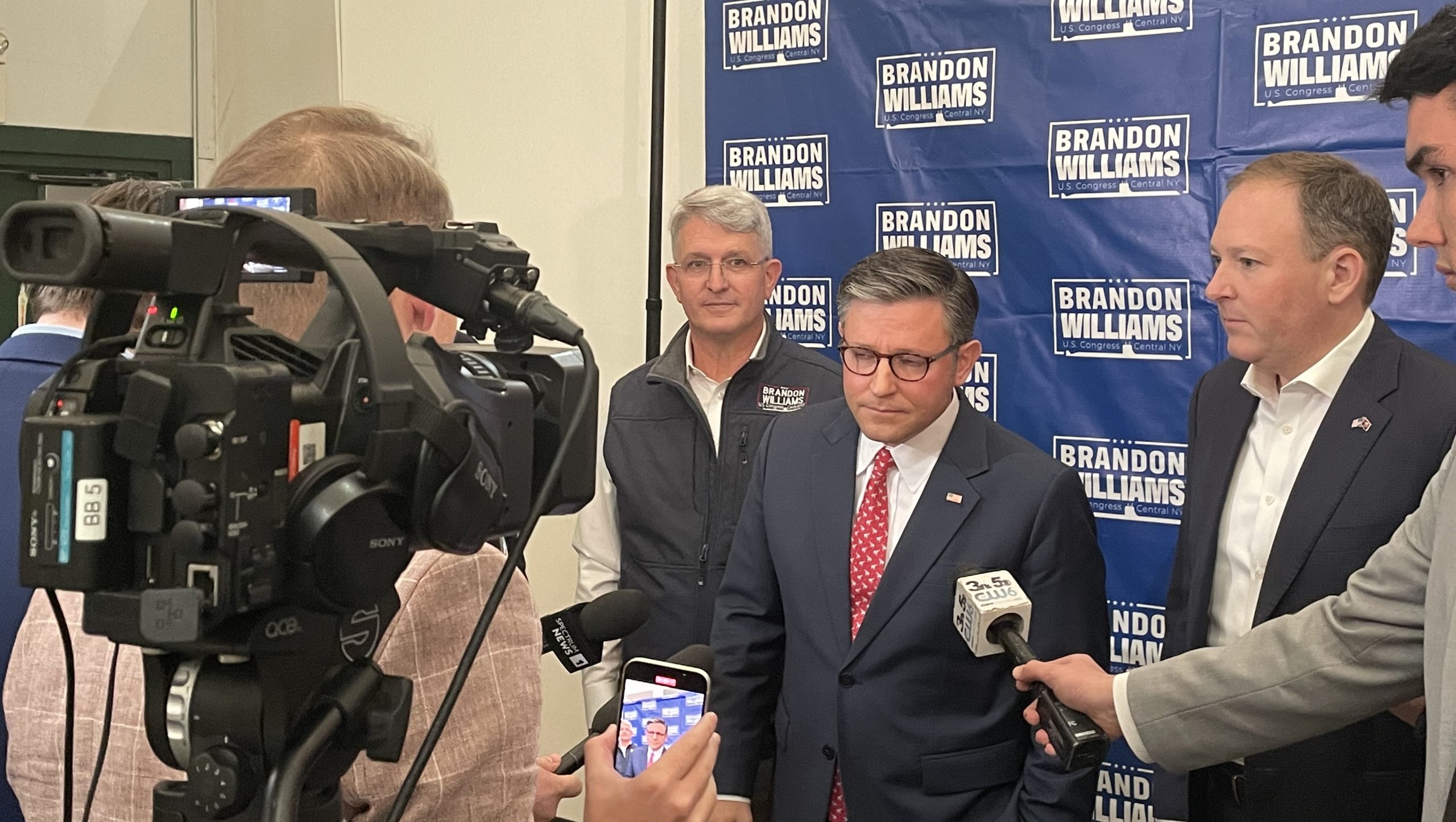 Speaker of the house Mike Johnson talks to TV reporters while standing next to Brandon Williams at an event in CNY.