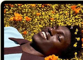 MacBook Neo, citrus color, screen shows a smiling person lying in a sunny field of flowers