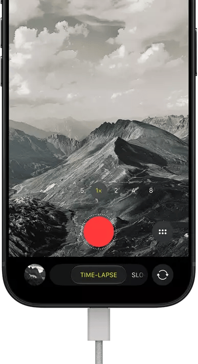 iPhone 18 Pro, front exterior, displaying a mountainous landscape, with the camera mode overlay in Time-Lapse mode, with power cable attached to charging port