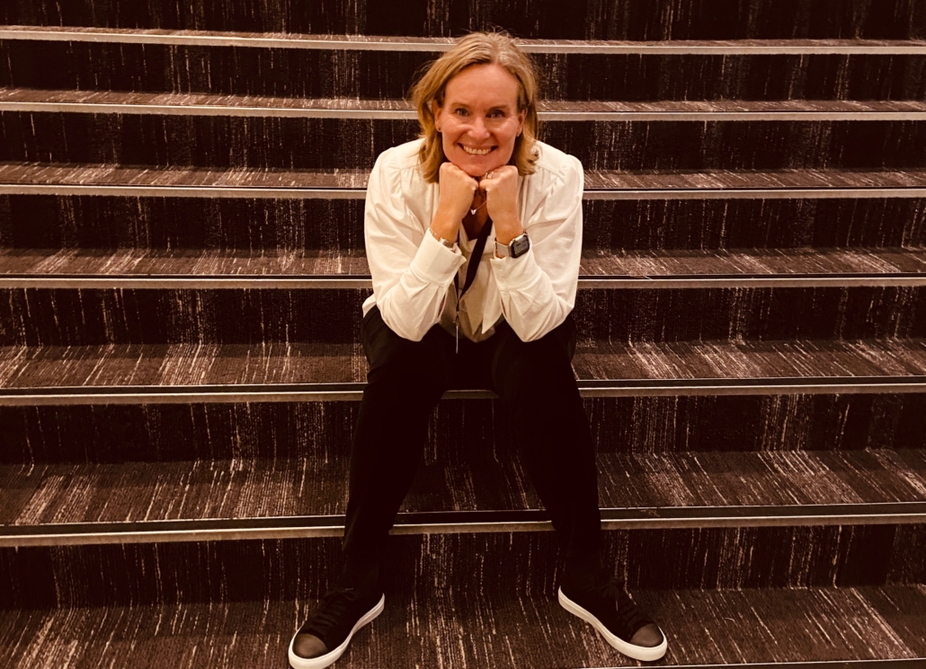 Lotta Broberg sitter i en trappa. Hon ler mot kameran och vilar ansiktet i sina händer.