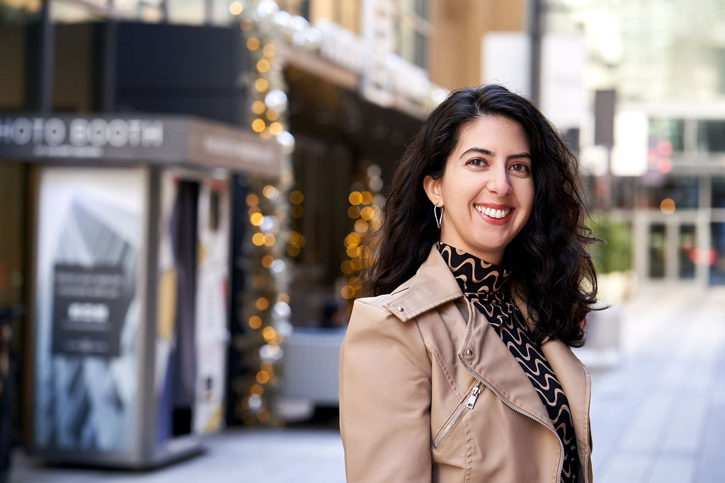 Headshot of Catarina Rivera, light-skinned Latine woman with long brown wavy hair, hoop earrings, a black and beige wavy striped top, and a beige jacket smiling at the camera and angling her body to the right. Behind her is a blurred background of a street with shops and a photo booth.