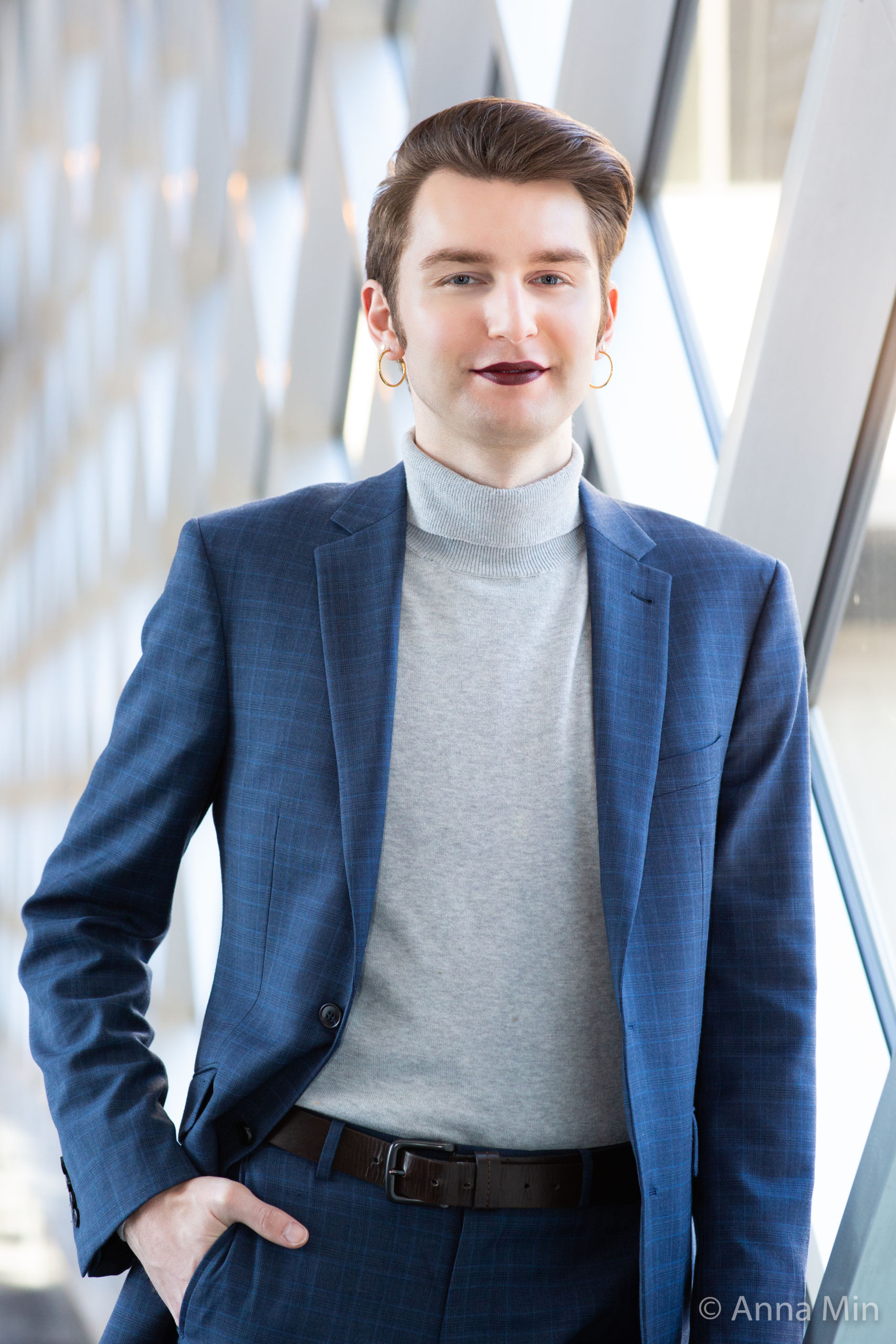 Headshot of Nick Alm, a White nonbinary person with short light brown hair, golden hoop earrings, eyeliner, sparkles, purplish-red lips, grey turtleneck, and plaid blue suit looking at the camera leaning against a white wall covered with diagonal windows with their hand in their pocket.