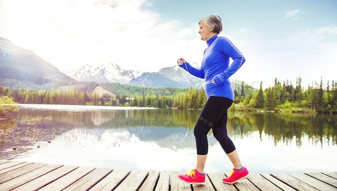 Older woman with red shoes walking on wooden path with lake and mountains in background