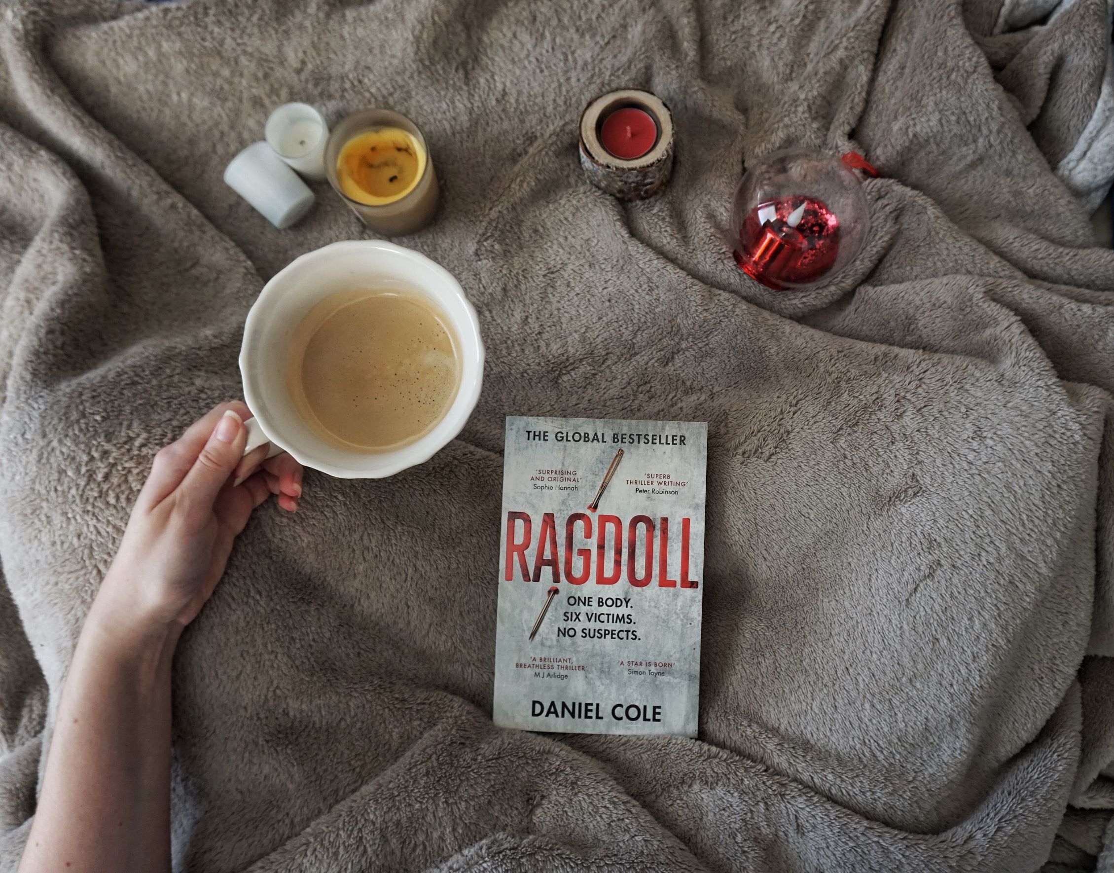 book on beige blanket with candles strewn around and a hand holding a cup of coffee