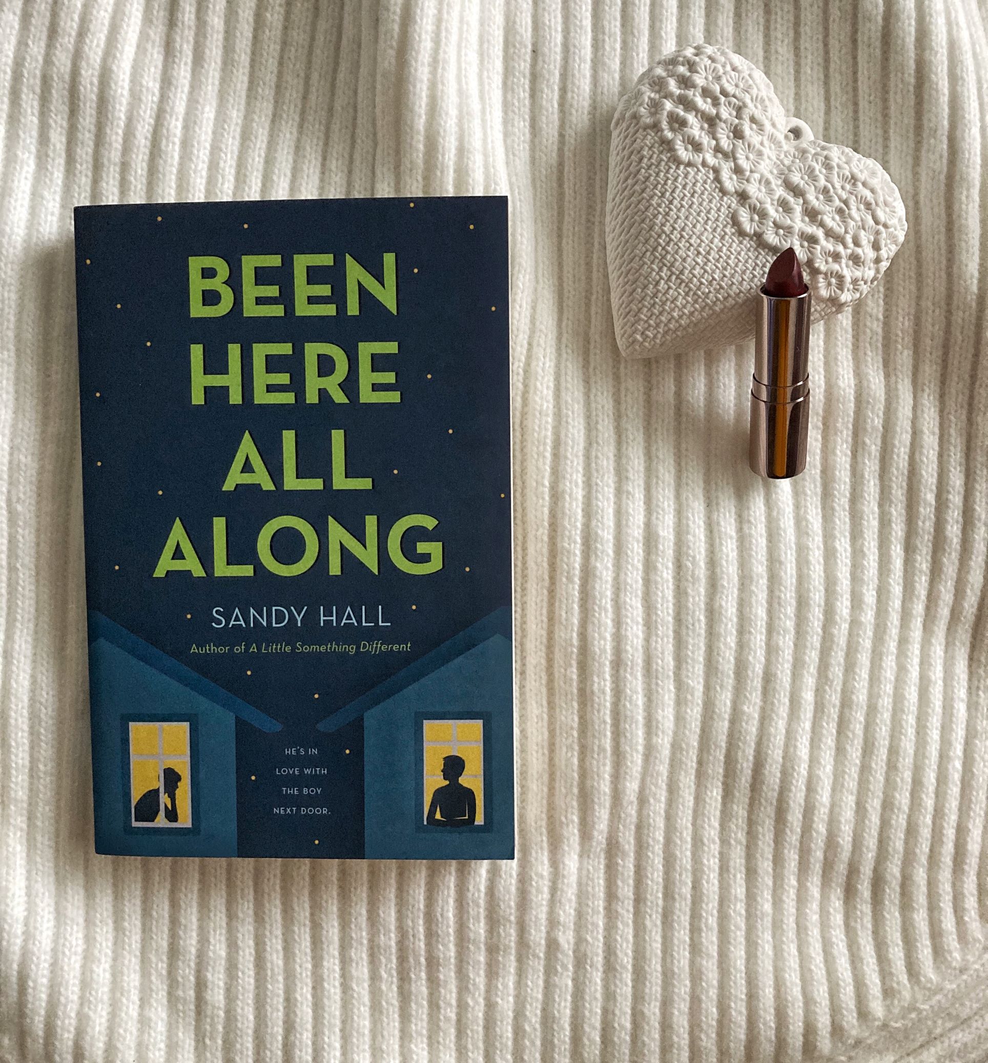 Flatlay of book 'Been Here All Along' on white striped background and red lipstick leaning on white ceramic heart