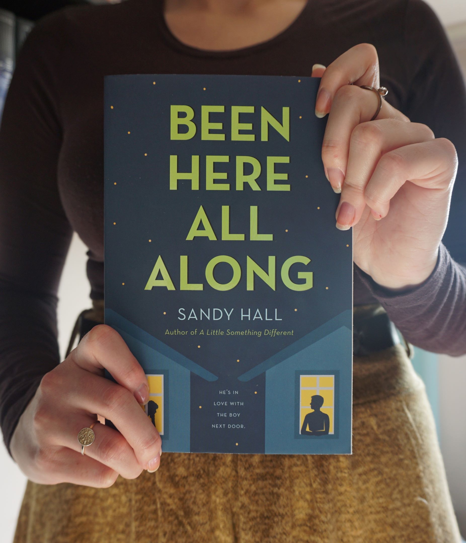girl holds copy of 'Been Here All Along' by Sandy Hall, dressed in brown long sleeved shirt and ocher skirt