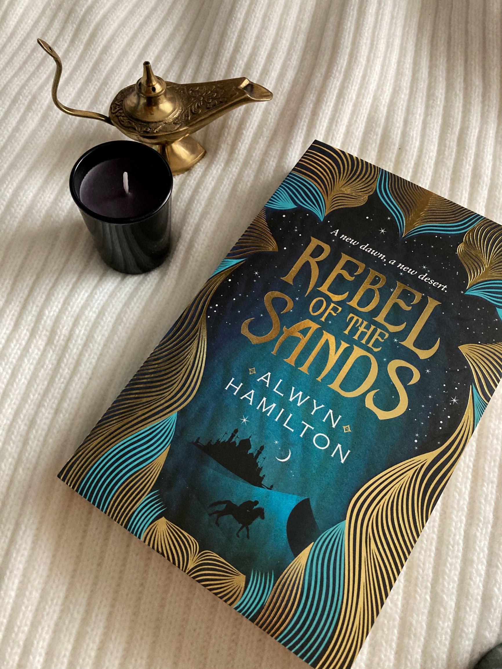 book review: Rebel of the Sands, book on white striped background next to dark candle and golden lamp