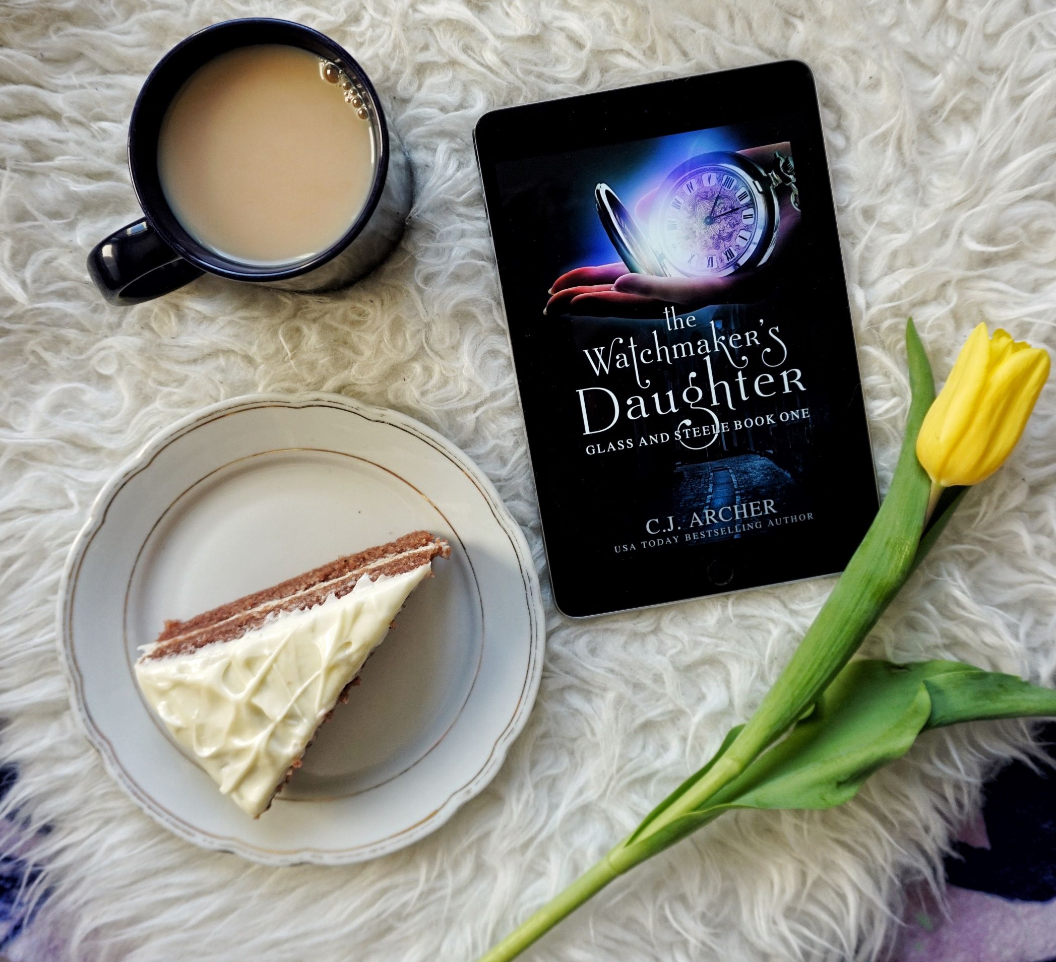 Flatlay shot of The Watchmaker's Daughter (E-book) on white fur with a yellow tulip ocerlapping it and a white plate with red velvet cake and white frosting next to it and a blue mug filled with sweetened black tea