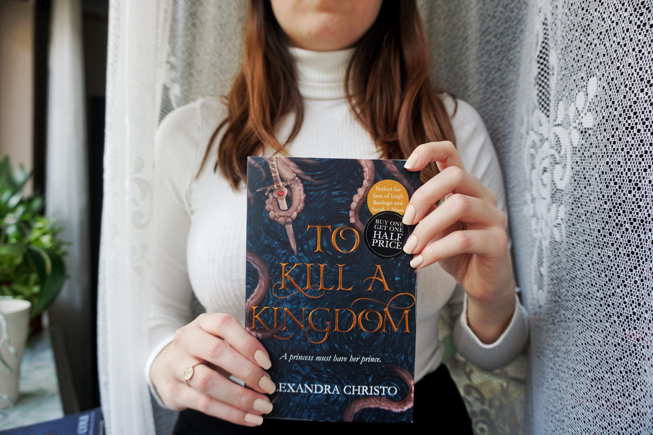 A brunet girl, dressed in a white striped, long-sleeved turtle neck, holding a paperback copy of To Kill A Kingdom by Alexandra Christo in both hands.
