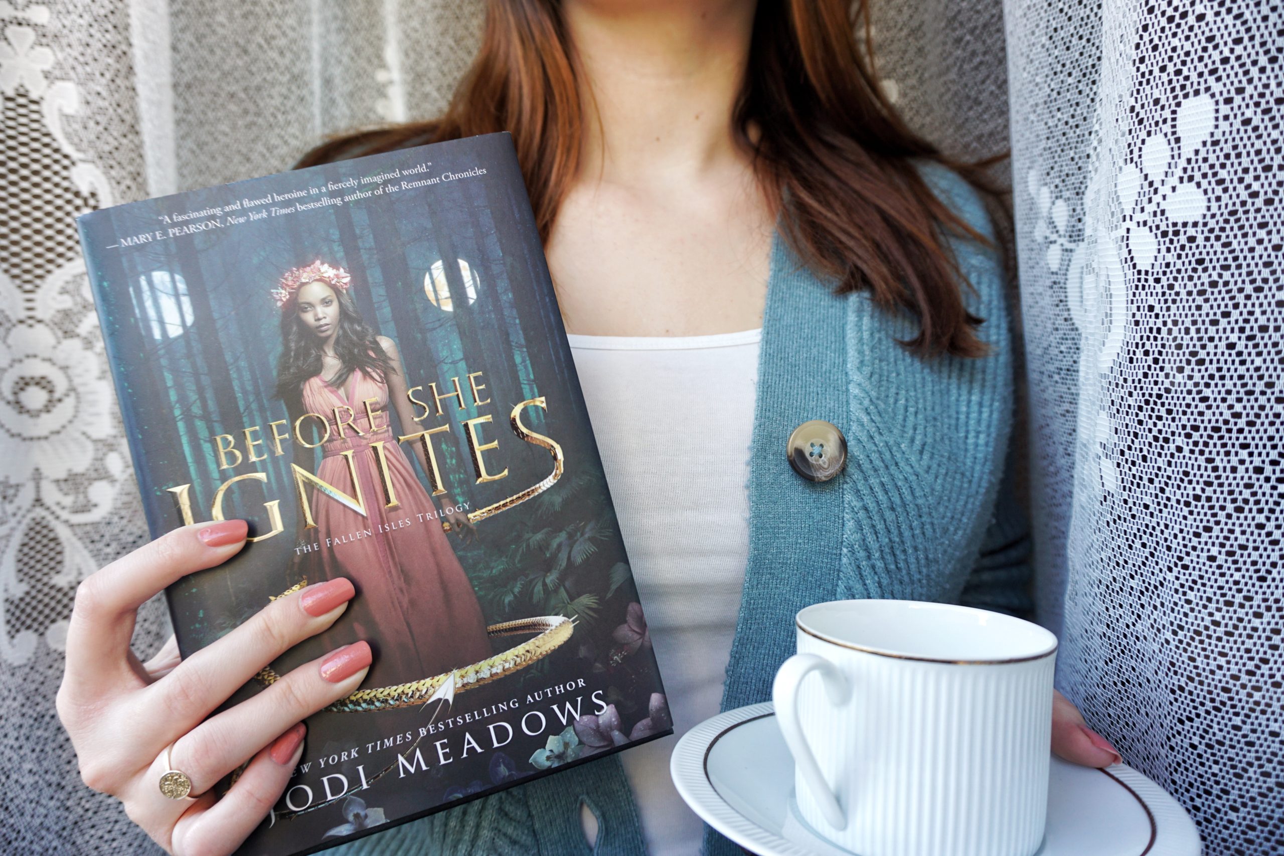 A brown haired girl, dressed in a blue cardigan and white top, holding a white tea cup on a saucer in one hand and a hardback copy of Before She Ignites by Jodi Meadows in the other. White embroidery in the background.