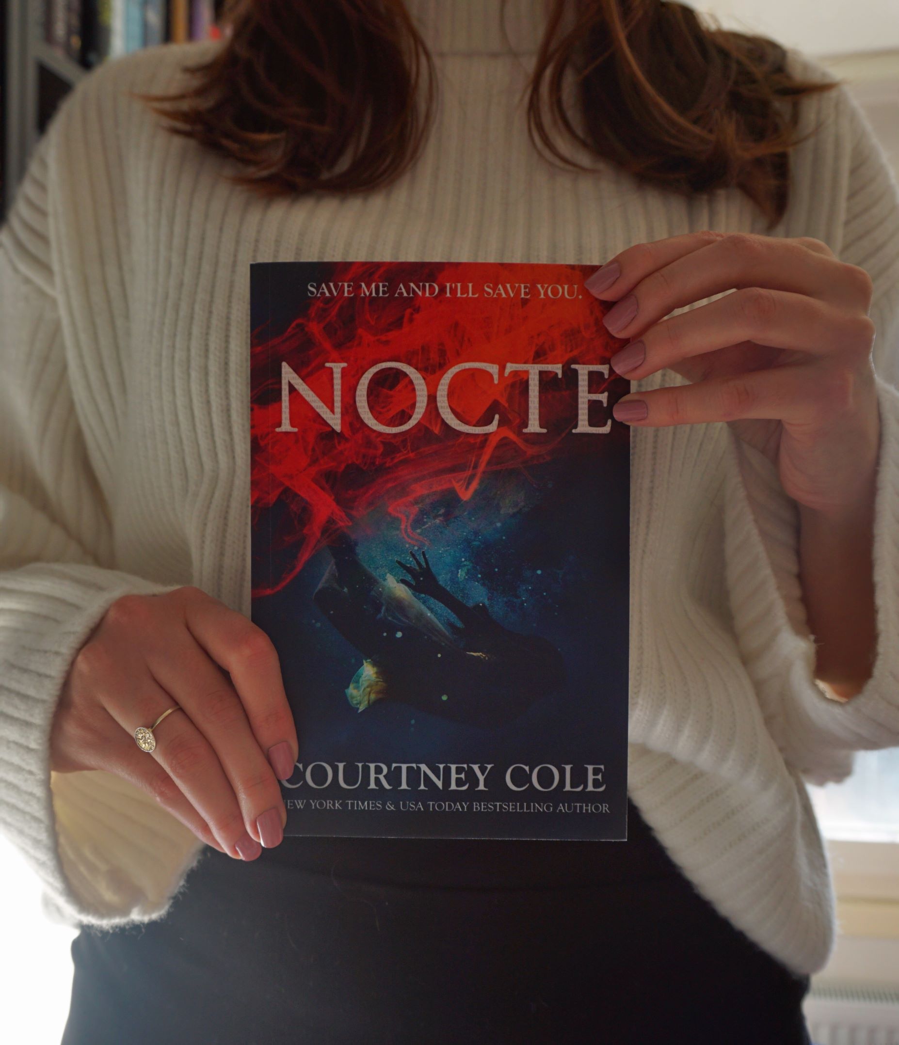 girl, dressed in dark blue skirt and white turtle neck jumper, holding a copy of Nocte by Courtney Cole, facing the camera