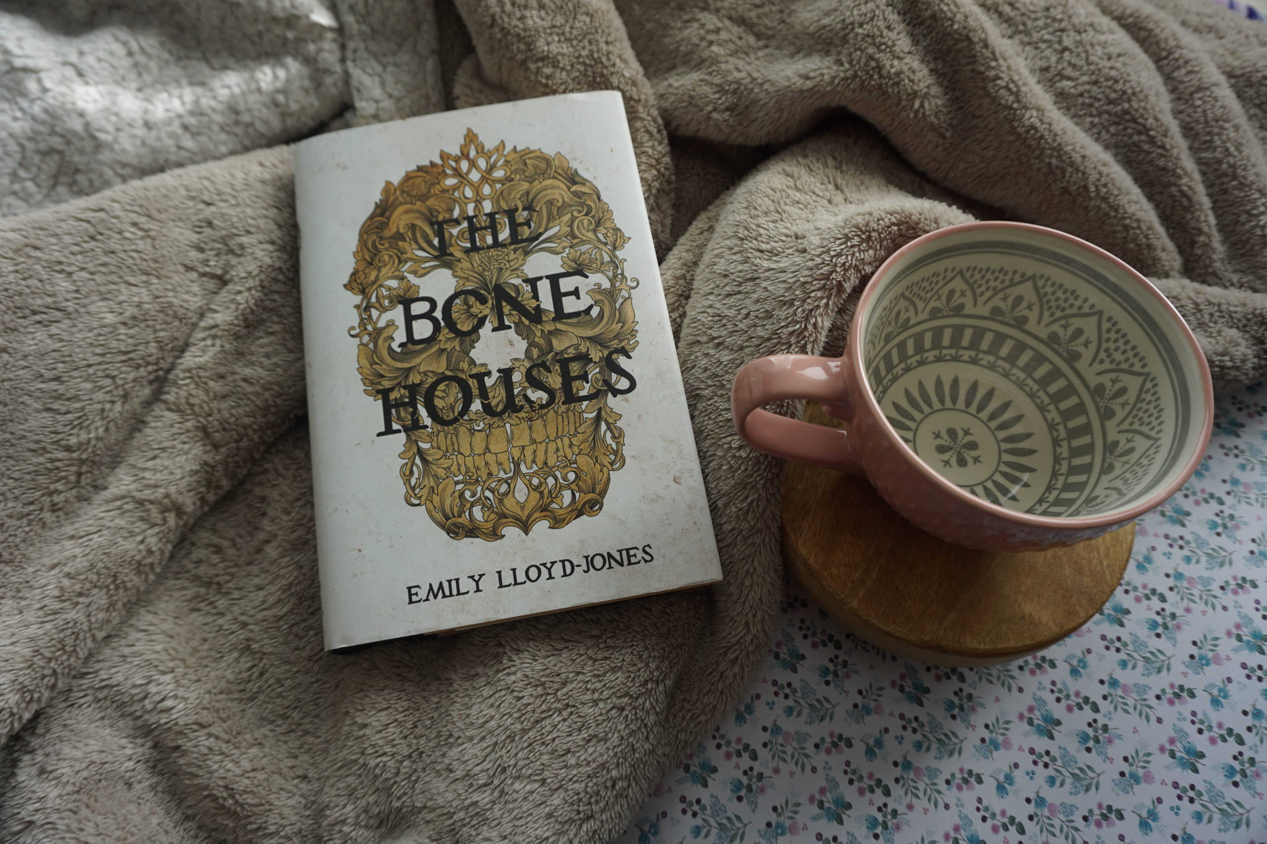 A hardcover copy of The Bone Houses lying on a beige blanket next to a baby pink mug resting on a wooden pedestal.