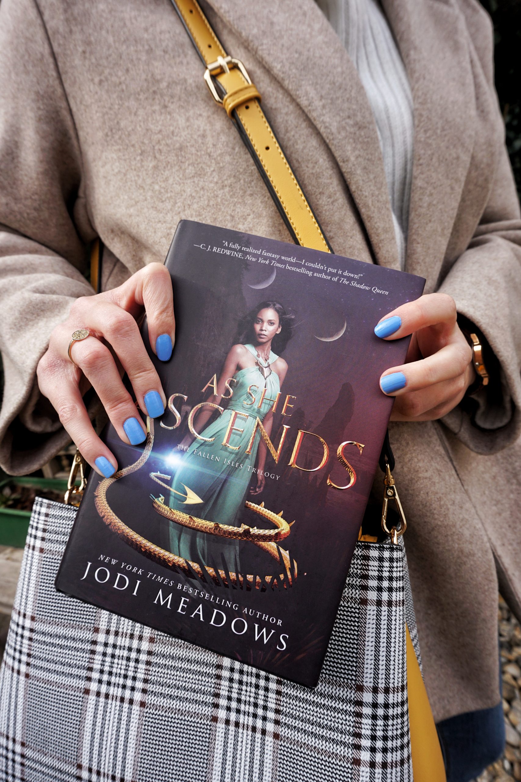 Picture of a girl, dressed in white shirt, blue jeans and beige coat, pulling a hardback copy of As She Ascends by Jodi Meadows out of her yellow, black and white striped bag.