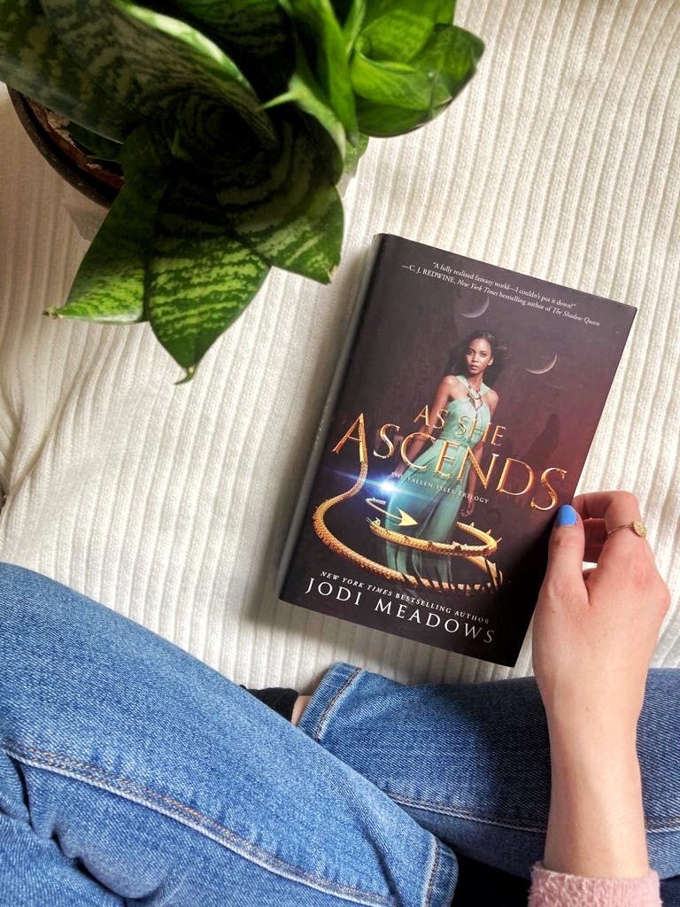 A flatlay shot from above. A hardback copy of As She Ascends by Jodi Meadows on a white striped background. Around it, a plant and someone wearing blue jeans, sitting cross-legged before it and touching the book.