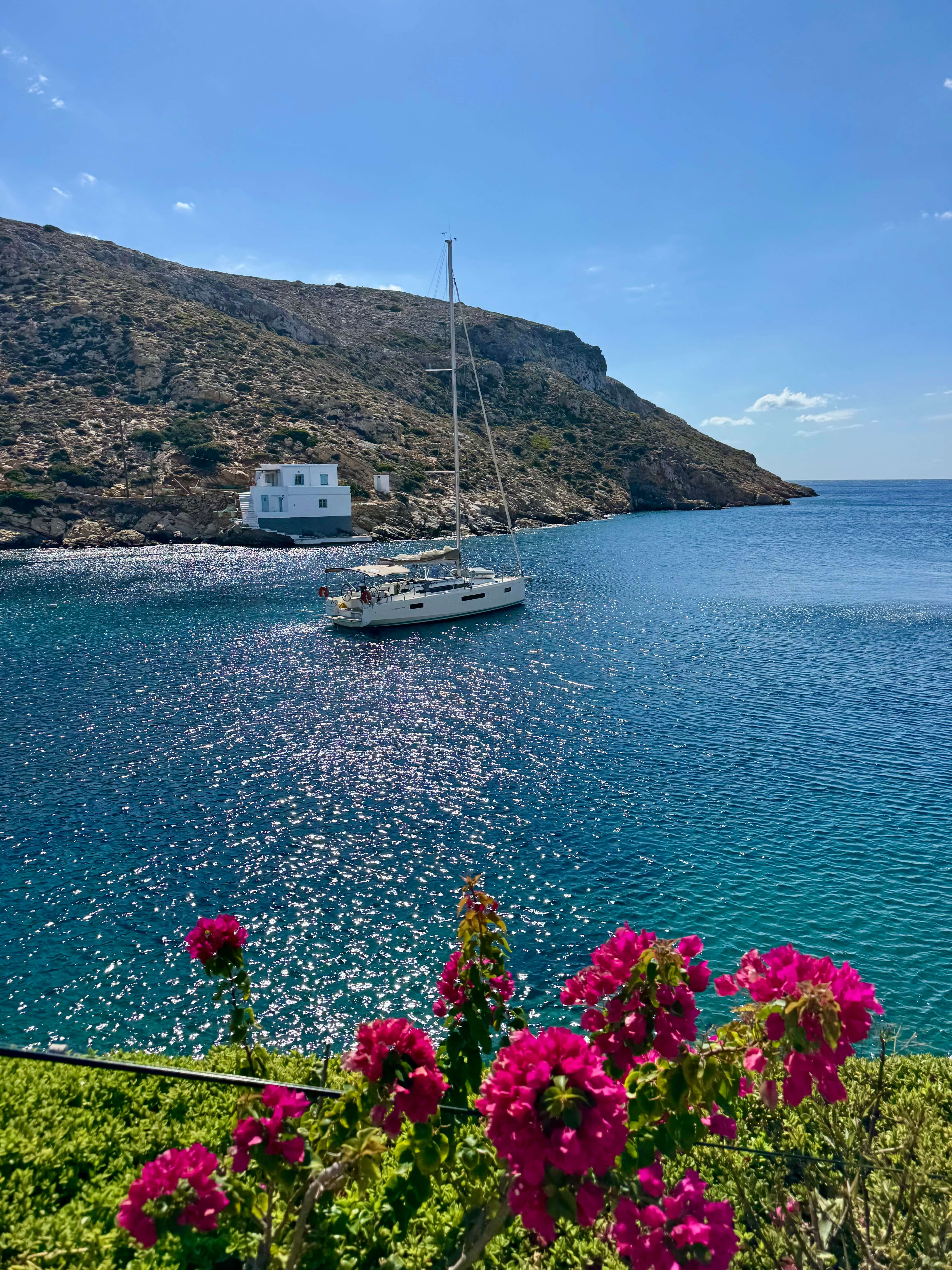 Sailing yacht anchored in a sheltered Cyclades cove, with pink flowers in the foreground.