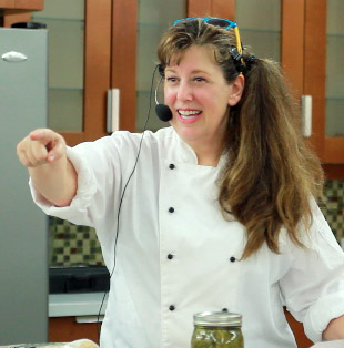 Chef Marian Meyers wears a white chef's jacket and points toward someone. She has a headset microphone on, with the mic beside her face. She wears a long side ponytail, and blue and yellow sunglasses on top of her head.